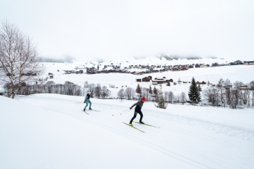 Zwei Personen beim Skaten auf einer Loipe vor einem verschneiten Bergdorf in Disentis