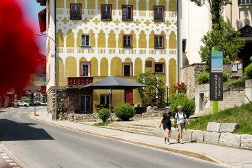 Zwei Wanderinnen gehen an einem historischen, gelb verzierten Haus in Savognin vorbei. Daneben weist ein Schild auf den Naturpark hin.