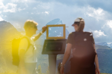 Zwei Wanderinnen betrachten das Bergpanorama durch einen gelben Kunstrahmen entlang der Senda Segantini.