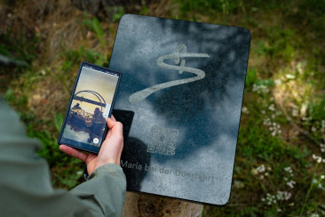 Eine Hand hält ein Smartphone vor eine Informationstafel mit QR-Code. Auf dem Bildschirm erscheint ein historisches Bild zur Geschichte der Überfahrt.