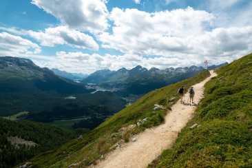 Zwei Wandernde steigen auf einem Bergpfad bergauf. Der Blick reicht über den grünen Engadiner Talboden mit mehreren Seen bis zu den Gipfeln.